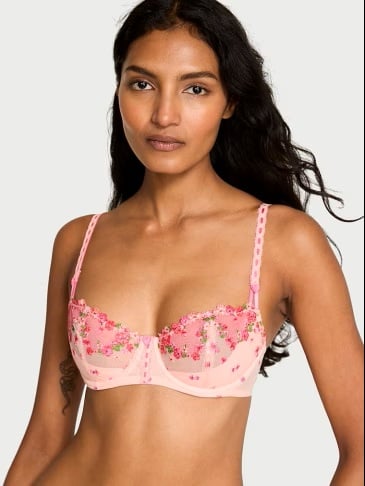 Woman posing in a Victoria's Secret pink unlined bra with flower details on a light grey background.